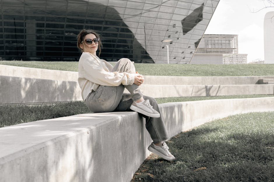 A stylish woman sits on concrete ledge near contemporary building, enjoying a sunny day.