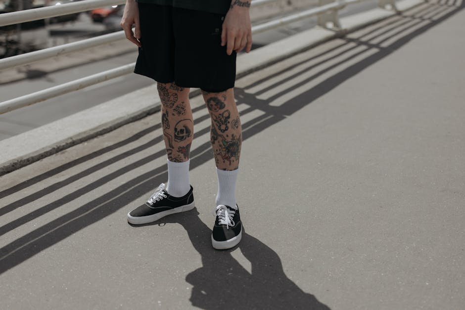 A person with tattooed legs standing outdoors in black shorts and sneakers, casting shadows on pavement.