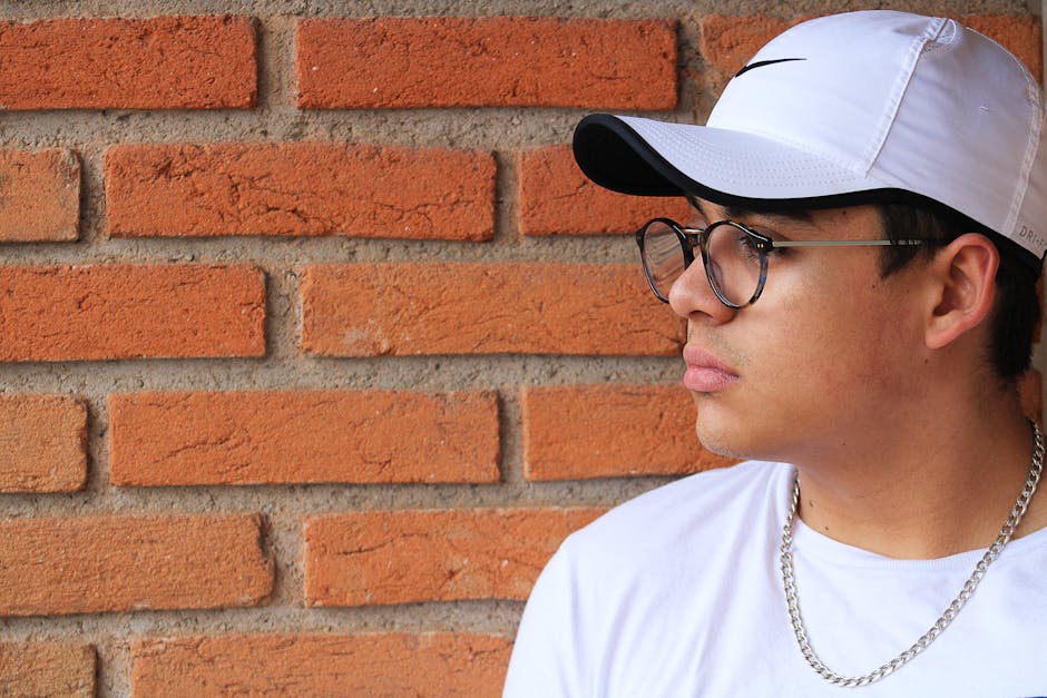 Profile of a young man wearing a white cap and glasses against a brick wall background.