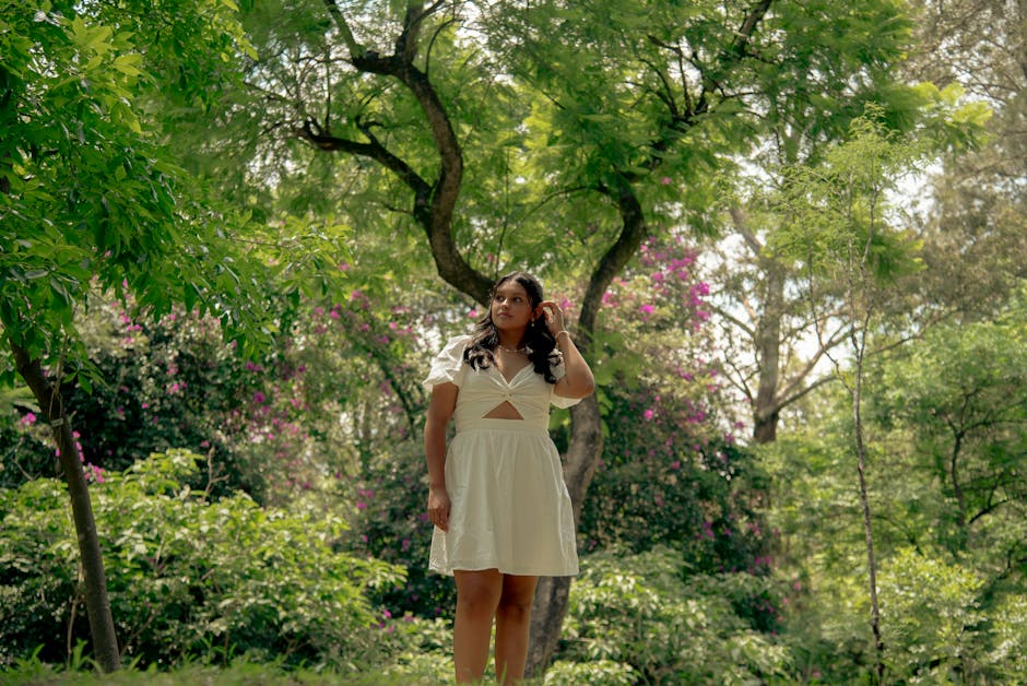 A woman in a white dress stands amidst lush greenery and vibrant flowers during the day.