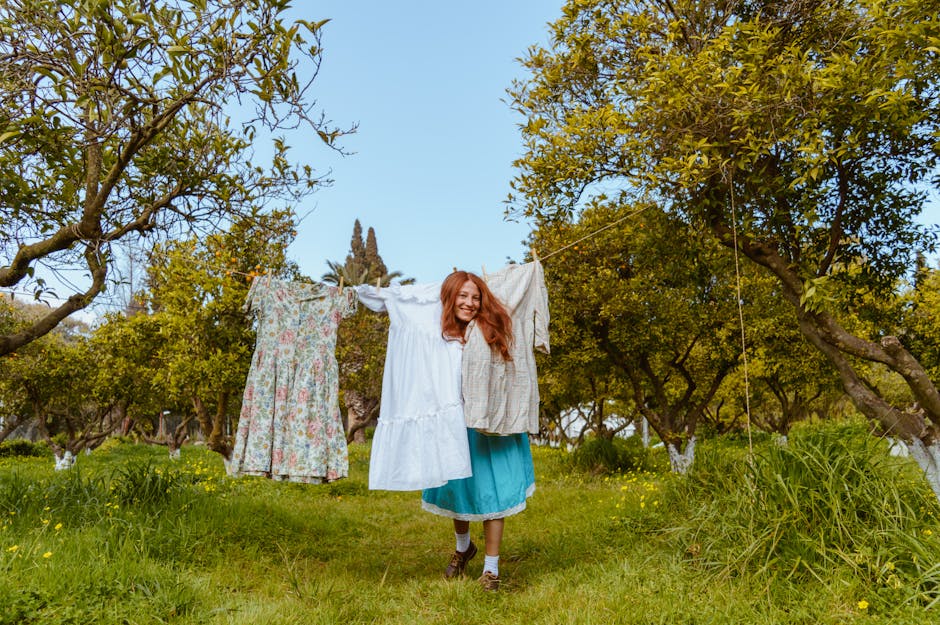 Smiling woman hanging dresses on a clothesline in a sunny orchard surrounded by green grass and trees.